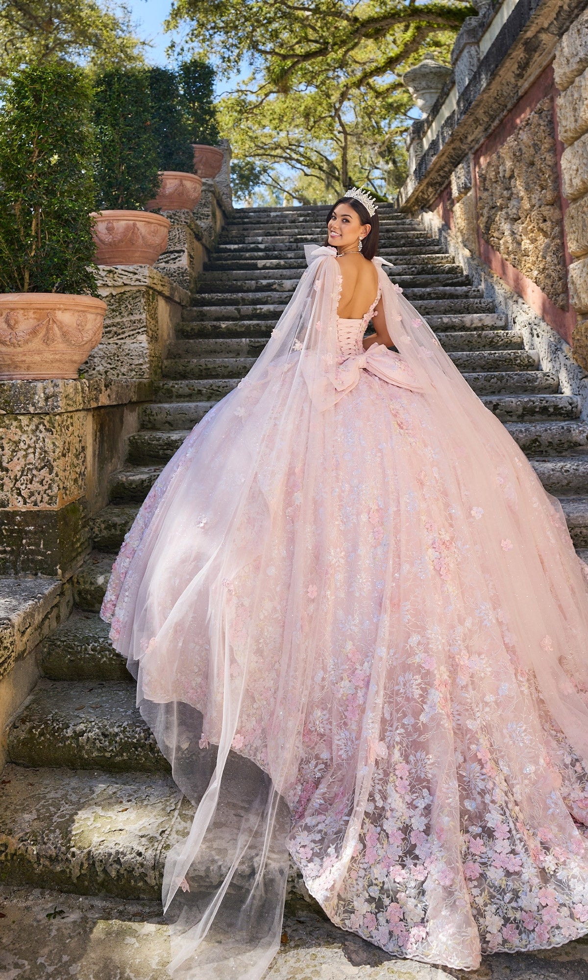 Wearing the Princesa PR30135, a pale pink 3D Floral Quinceanera Dress with a corset bodice and long, sheer train, a woman stands on outdoor stone steps looking back over her shoulder.